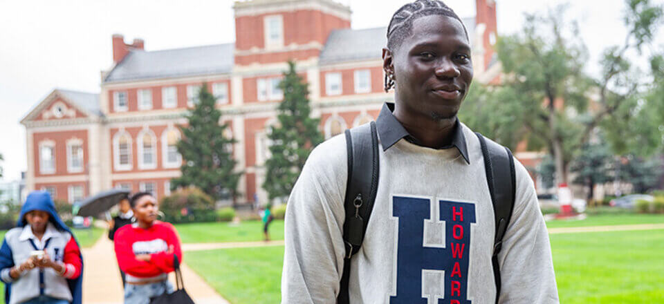 Student wearing a large pale sweater with the Howard H featured on the front walking through campus with a bookbag and smiling. More students in Howard sweaters carry backpacks on the path behind him, with Founders Library in the background.