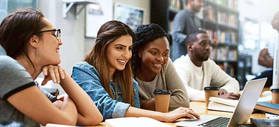 Three women seated at a library table looking at a laptop. More students are seated at the table in the background or standing near bookshelves.