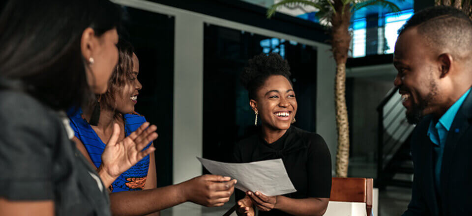 Two women in a nice office seated a table smiling at their colleague who holds a page. A man is seated nearby, also smiling at the woman.