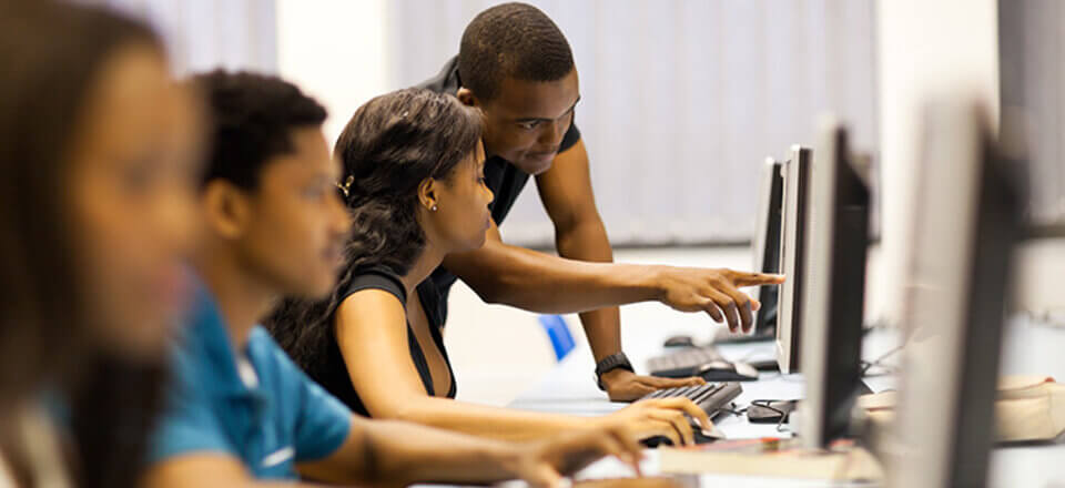 A view of down a row of students seated at a long table using computers as a professor point to one screen and explains something to a student.